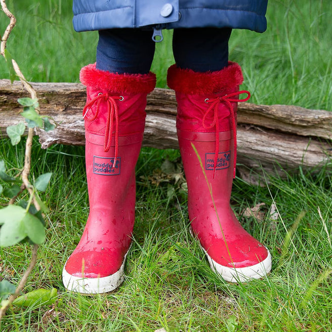 A pair of red 'PuddleFlex Fleece Lined Wellies' stand in grass. The boots have fluffy red tops and white soles. A small log is behind them.